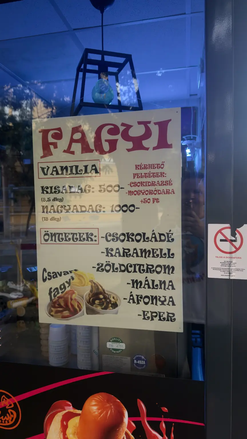 Kézzel írt vaníliafagylalt-árlista két adagmérettel, hat öntettel és két feláras feltéttel egy üzlet kirakatában