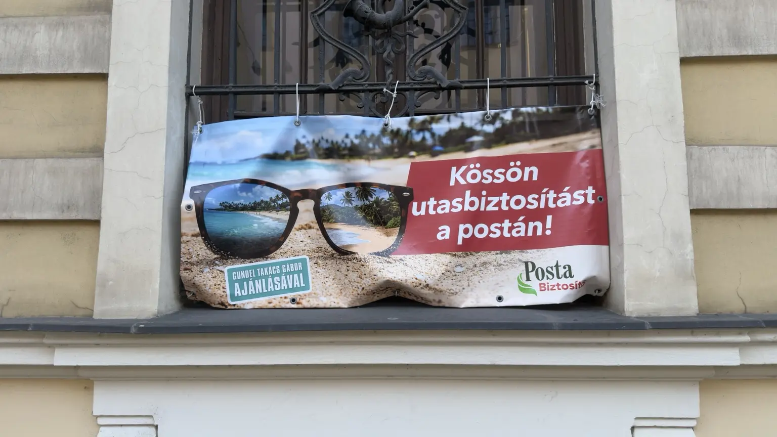 Posta Biztosító ablakra szerelt utasbiztosítási hirdetése tengerparttal, napszemüveggel és Gundel Takács Gábor ajánlására utaló jelvénnyel