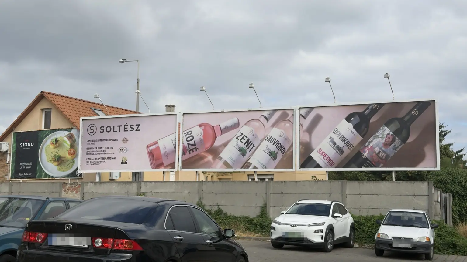 Három egymás melletti Soltész óriásplakát borospalackokkal, díjakkal és egységes rózsaszín háttérrel