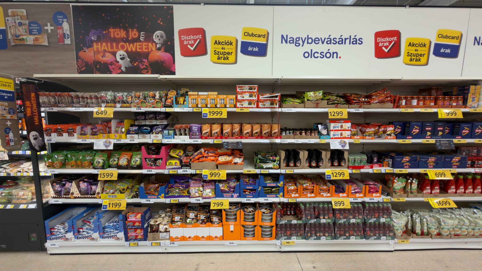 Tesco polcsor Tök jó Halloween felirattal, több márka szezonális édességeivel és a jobb oldalon további élelmiszerekkel